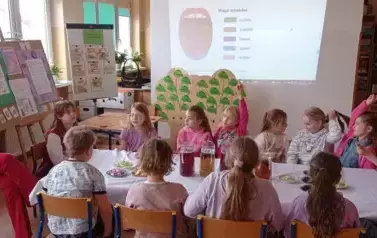 Na zdjęciu widać grupę dzieci siedzących przy stole, na kt&oacute;rym znajdują się owoce i napoje. Dzieci uczestniczą w lekcji, a na tablicy interaktywnej wyświetlona jest prezentacja dotycząca smak&oacute;w.