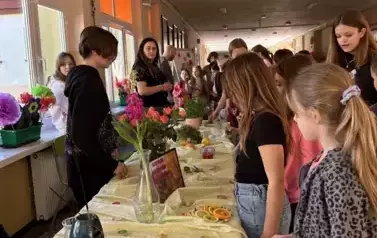 Na zdjęciu widać grupę dzieci zgromadzonych wok&oacute;ł stołu w szkolnym korytarzu, na kt&oacute;rym znajdują się r&oacute;żnorodne przedmioty rozwijające zmysł węchu.