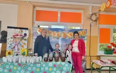 Na zdjęciu widać dwie kobiety i dwoje dzieci stojących za stołem przykrytym kolorowym obrusem, na kt&oacute;rym znajdują się szklanki i dzbanki z herbatą. W tle widoczne są okna z pomarańczowymi ramami i dekoracje ścienne.