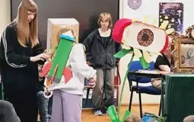 Na zdjęciu widać grupę szkolną, gdzie dzieci odgrywają przedstawienie. W centrum kadru stoi dw&oacute;jka dzieci w kostiumach, a za nimi, na tle tablicy, trzecie dziecko obserwuje scenę.